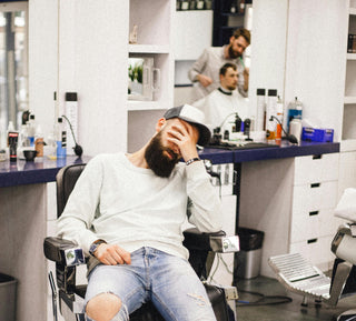 Man mit Vollbart beim Barbier auf dem Stuhl mit der Hand auf dem Gesicht